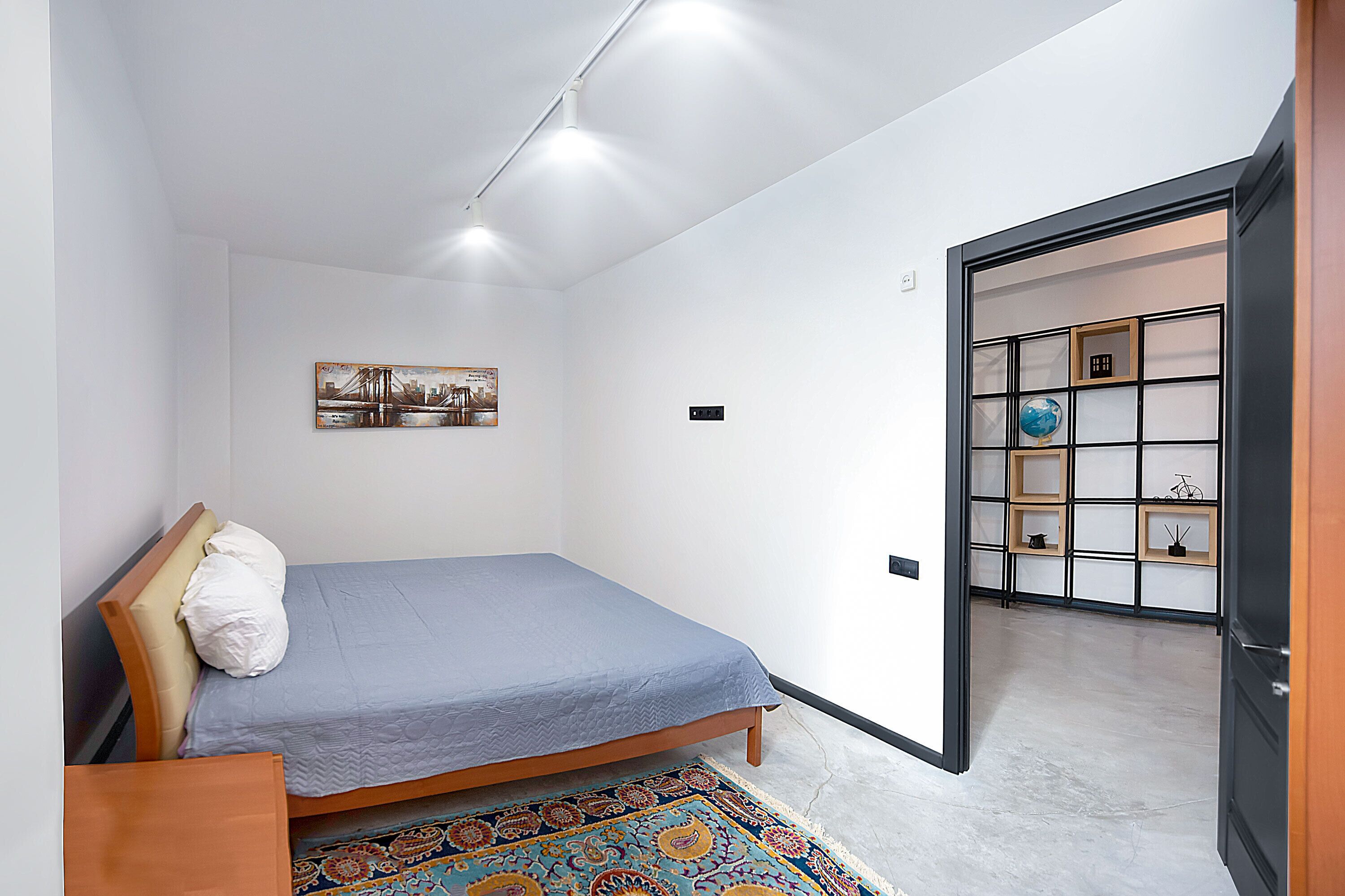 A bedroom with a bed, a headboard, a bedside table, a rug, a wall-mounted shelf with decorative items, and a door leading to another room.