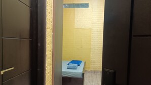Ein schmaler Flur führt zu einem kleinen Zimmer mit einem Bett, einem Holzschrank und einer Wand mit gelber Struktur.