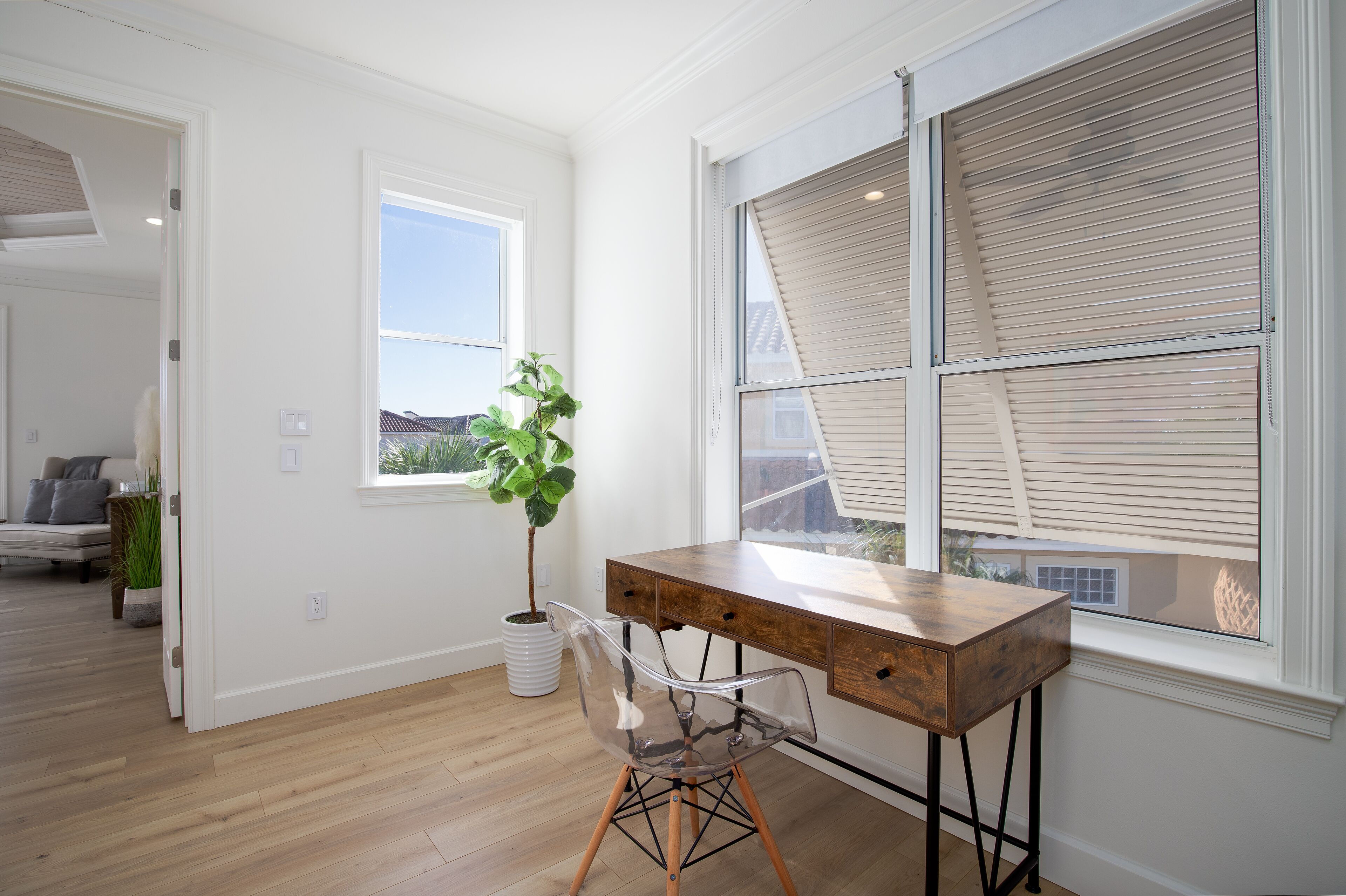 Une pièce avec un bureau en bois, une chaise transparente, une plante en pot et une fenêtre avec des stores.
