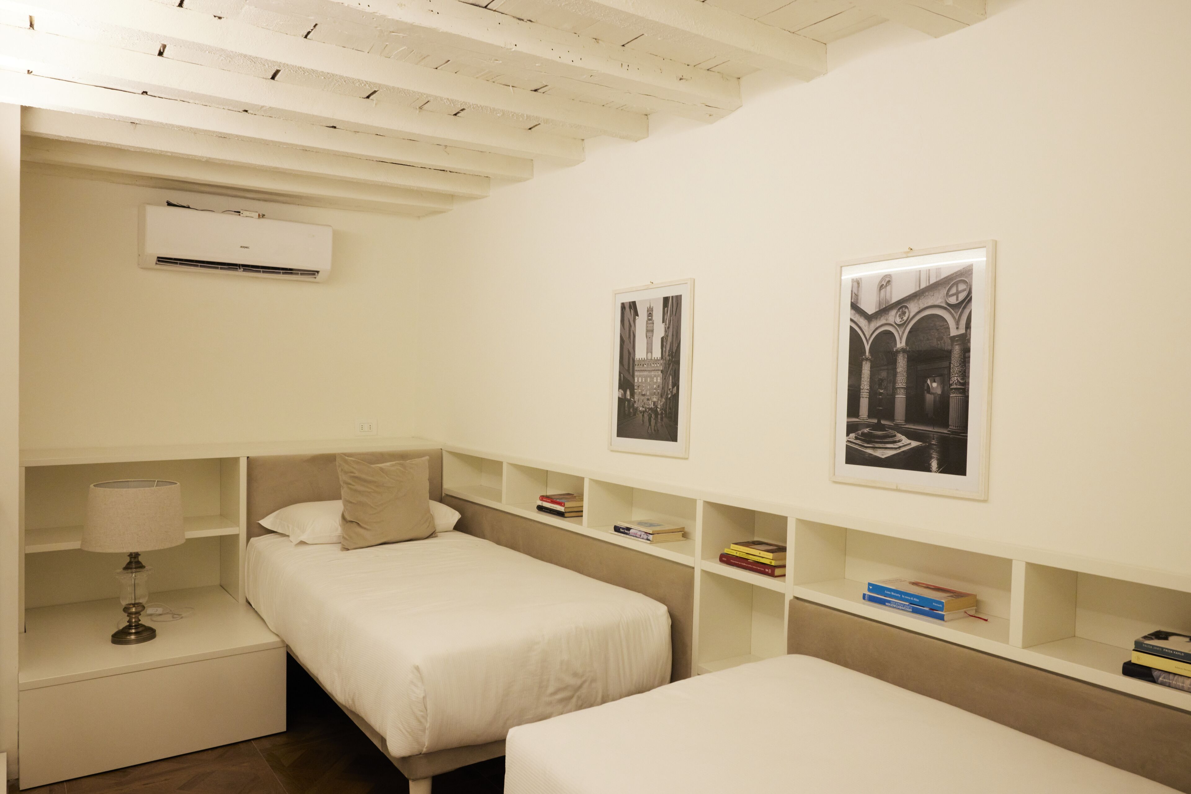 Een slaapkamer met twee bedden, een nachtkastje met een lamp en planken met boeken en ingelijste foto's.