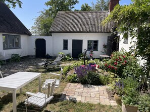 Une maison blanche avec un toit en tuiles, un jardin avec différentes plantes et du mobilier d’extérieur.