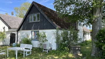 Une maison avec un toit en pente, une façade blanche et un petit jardin agrémenté d’un banc et d’un appareil de climatisation.