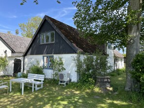 Une maison avec un toit en pente, une façade blanche et un petit jardin agrémenté d’un banc et d’un appareil de climatisation.