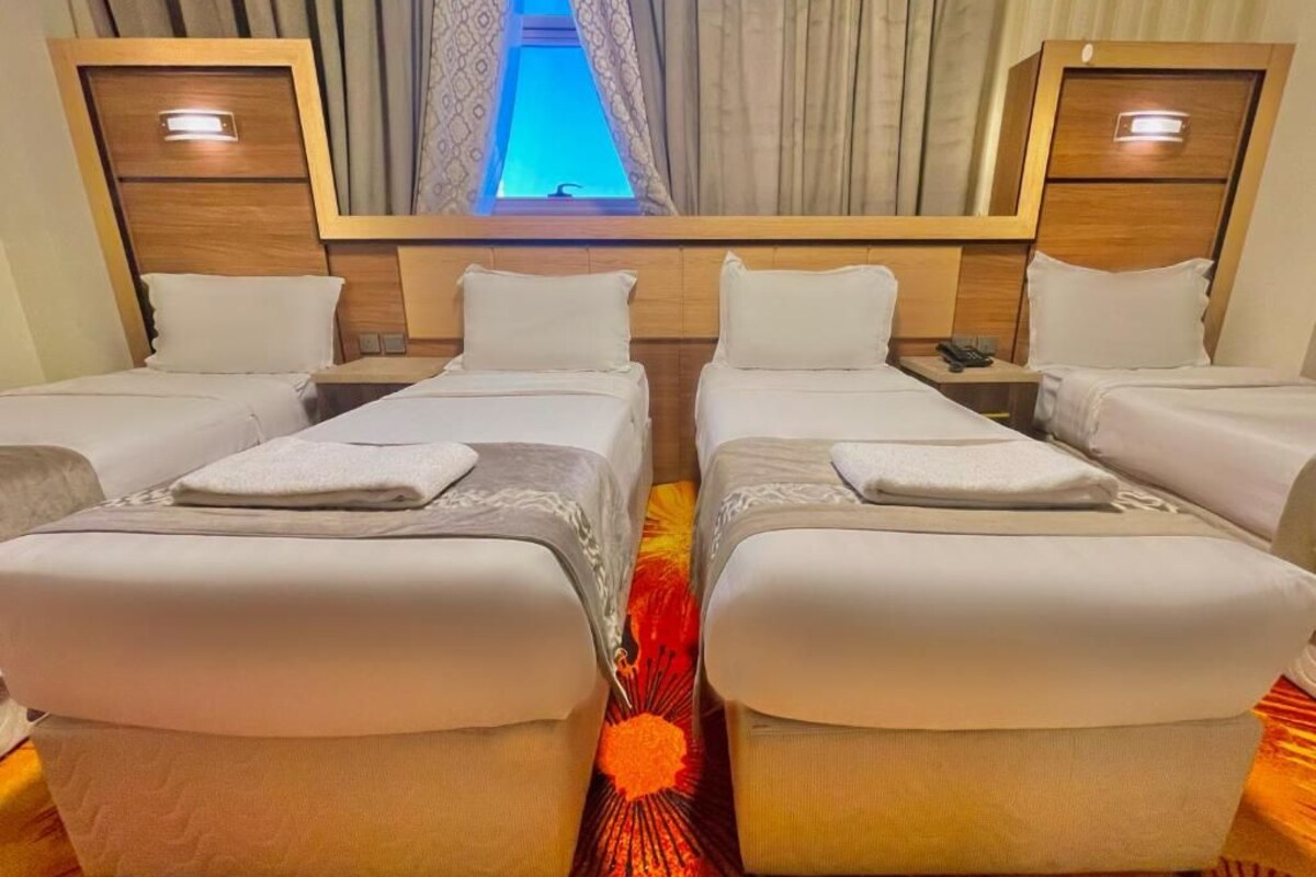 A hotel room with four single beds arranged in a row, a large mirror above the beds, and a window with curtains.