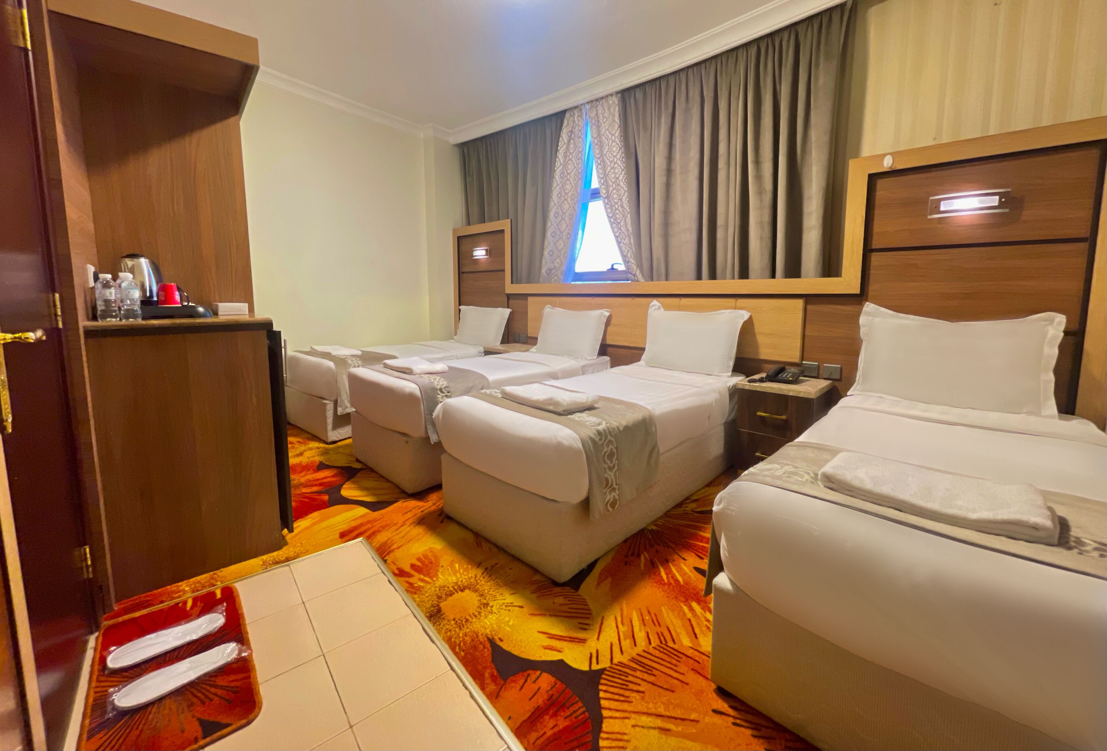 A hotel room with two single beds, a double bed, a wooden wardrobe, a desk with a telephone, and a window with curtains.
