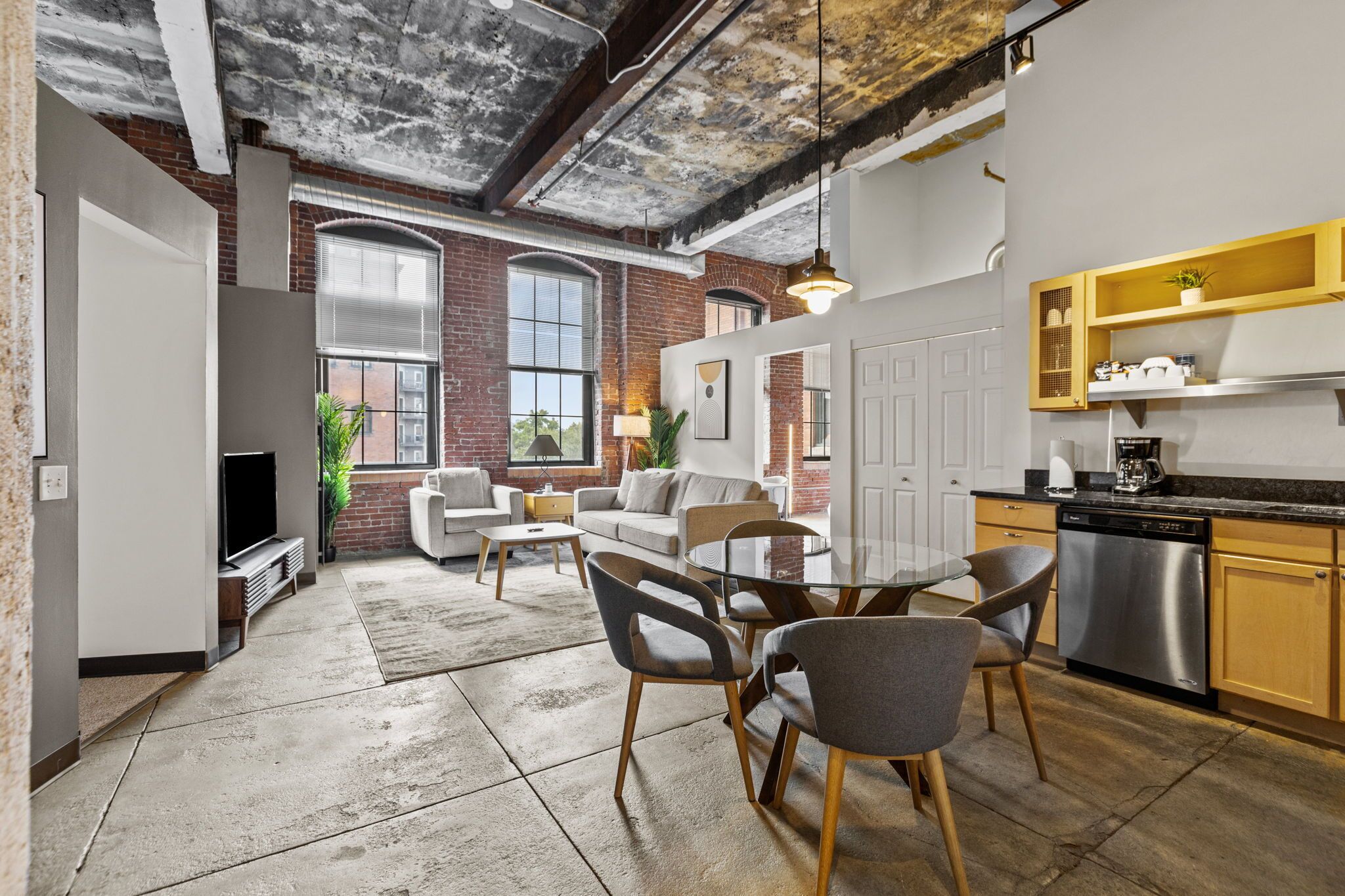 A modern kitchen with a dishwasher, a dining area with a round table and chairs, a living room with a sofa and a TV, and a brick wall with large windows.