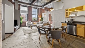 A modern kitchen with a dishwasher, a dining area with a round table and chairs, a living room with a sofa and a TV, and a brick wall with large windows. - Historic Loft Retreat With Pool (Pittsburgh)