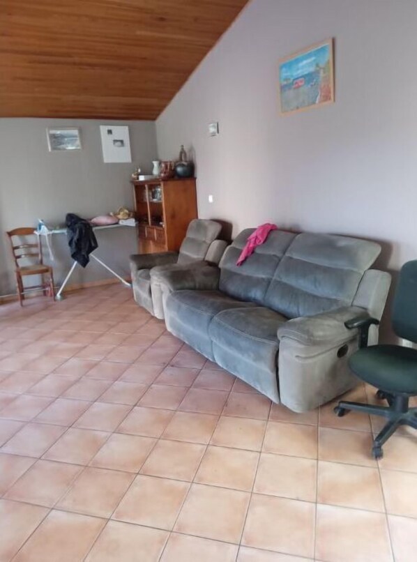 A living room with a grey sofa, a wooden chair, a tiled floor, and a painting on the wall.