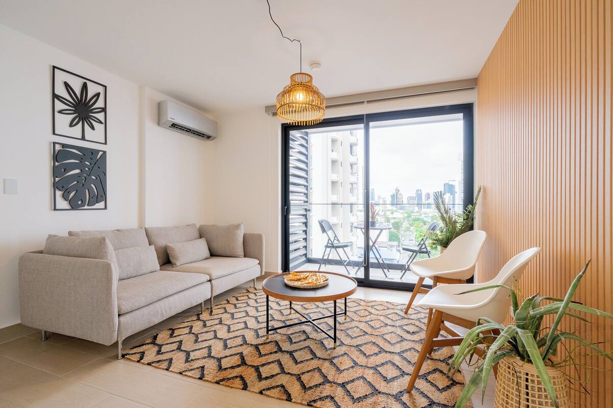A modern living room with a sofa, a round coffee table, a hanging light fixture, and a large window with a city view.