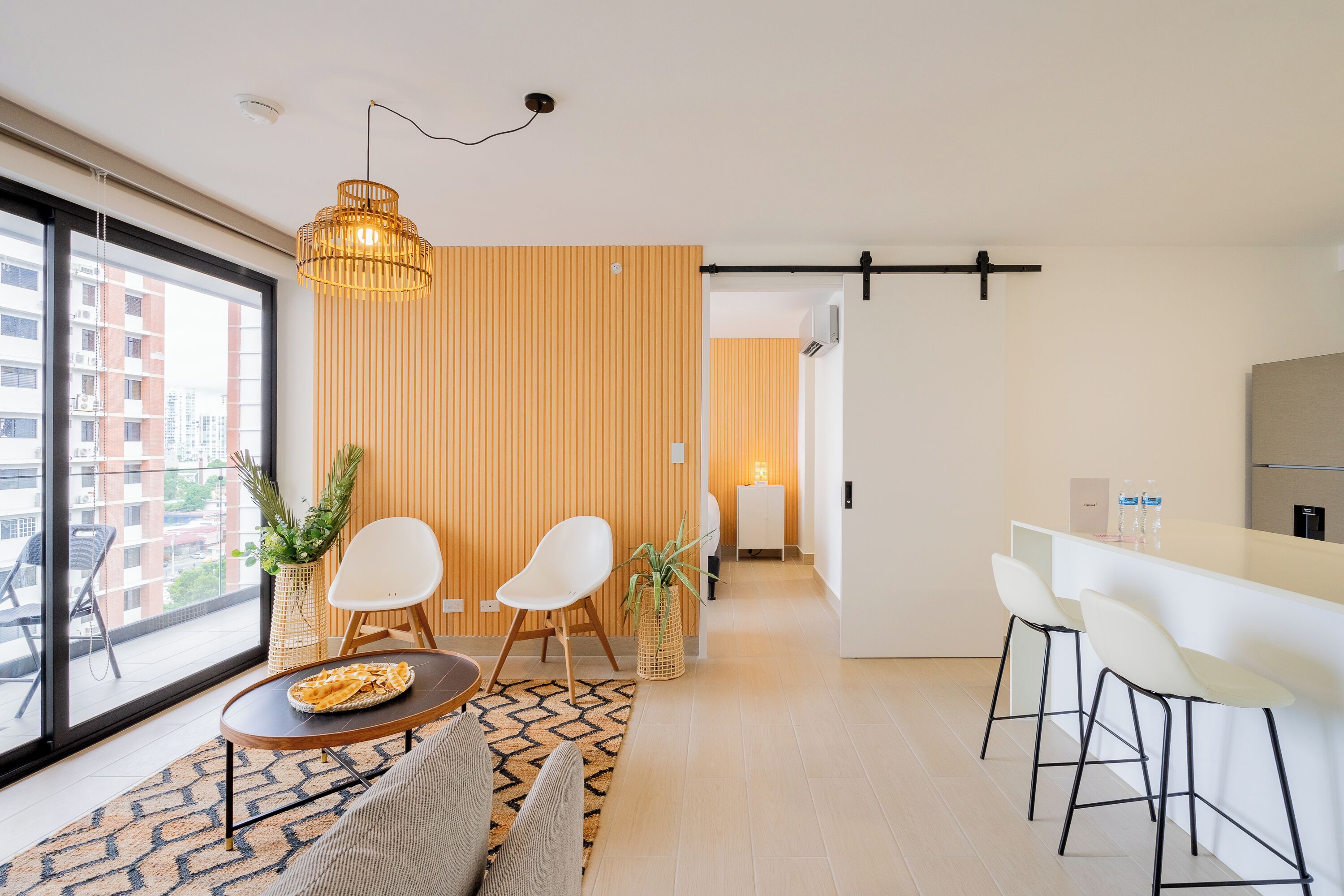 A modern living room with a sofa, a coffee table, and two chairs. There is a kitchen area with a bar and stools.