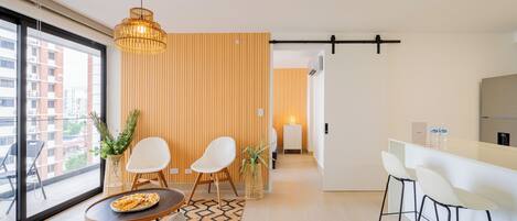A modern living room with a sofa, a coffee table, and two chairs. There is a kitchen area with a bar and stools.
