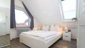 A bedroom with a bed, bedside tables, a radiator, a window with curtains, and a view of buildings outside.