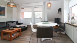 A modern living room with a black sofa, a wooden coffee table, a white dining table with four chairs, and a hanging pendant light.