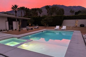 Ein rechteckiger Swimmingpool mit glasklar blauem Wasser, umgeben von einer gepflasterten Liegefläche. Vier weiße Liegestühle sind am Poolrand aufgereiht.