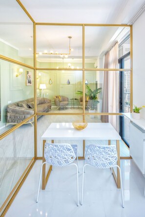 Uma sala de estar moderna com uma mesa branca, duas cadeiras estampadas, um sofá e uma divisória de vidro com molduras douradas.