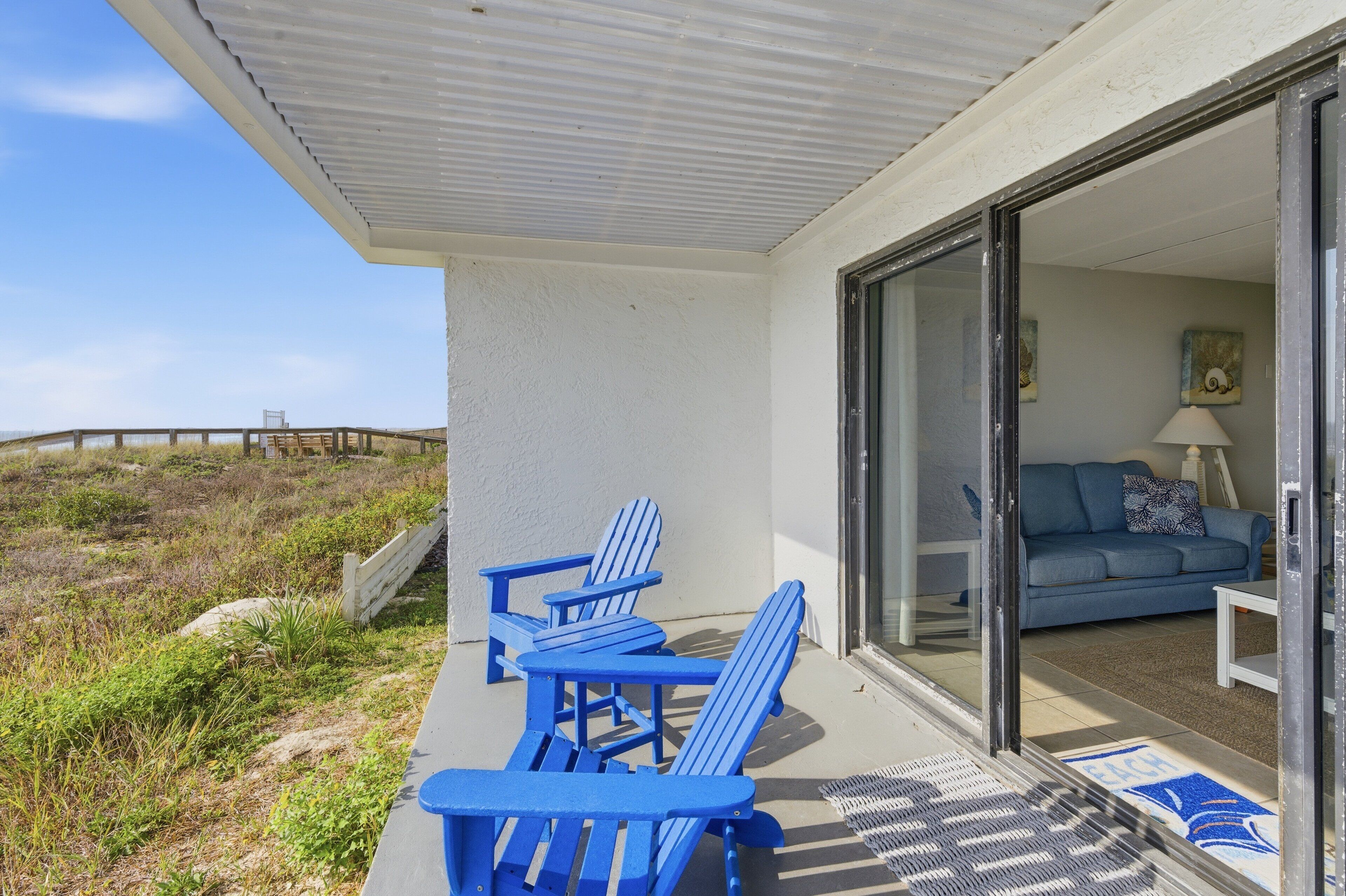 A covered outdoor seating area with blue Adirondack chairs, a glass door leading to a living room with a sofa and a lamp, and a view of the beach.