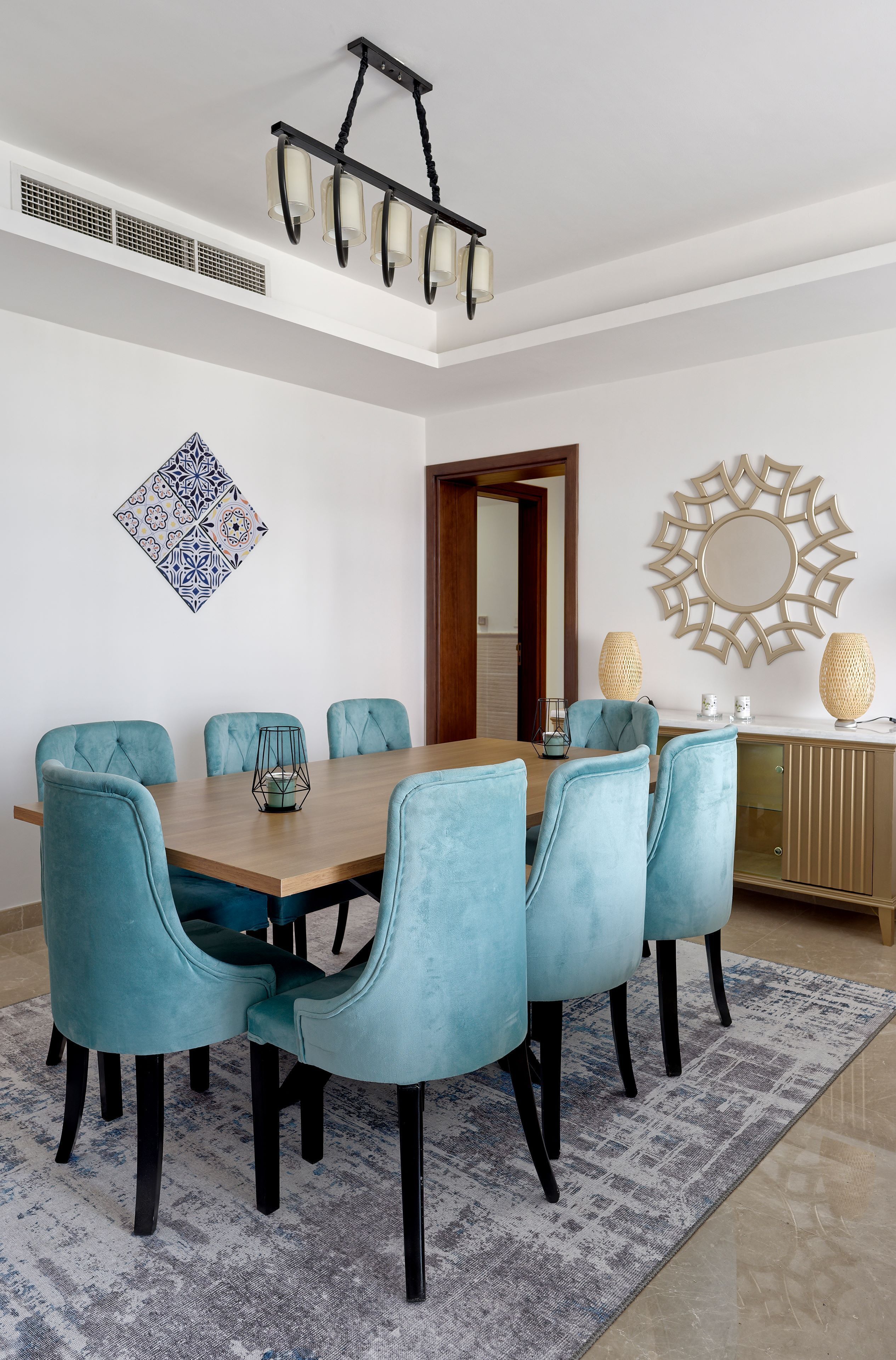 A dining area with a wooden table, six teal chairs, a patterned wall decoration, a decorative ceiling light, and a mirror on the wall.