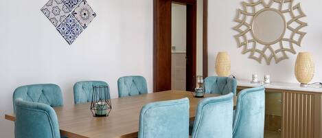 A dining area with a wooden table, six teal chairs, a patterned wall decoration, a decorative ceiling light, and a mirror on the wall.