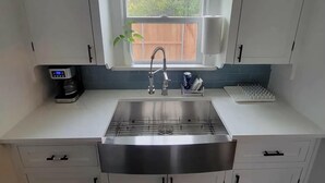 A modern kitchen with white cabinets, a stainless steel sink, a coffee maker, and a window with a view of trees. - Blanche's Bungalow (Graham)