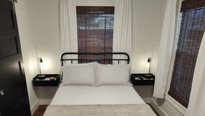 A bedroom with a bed, two bedside tables, a nightstand, a window with blinds, and a door. - Blanche's Bungalow (Graham)