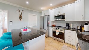 Una cocina moderna con armarios blancos, un refrigerador de acero inoxidable, un microondas, un horno y una cafetera. Hay dos sillas azules y una mesa pequeña con botellas.