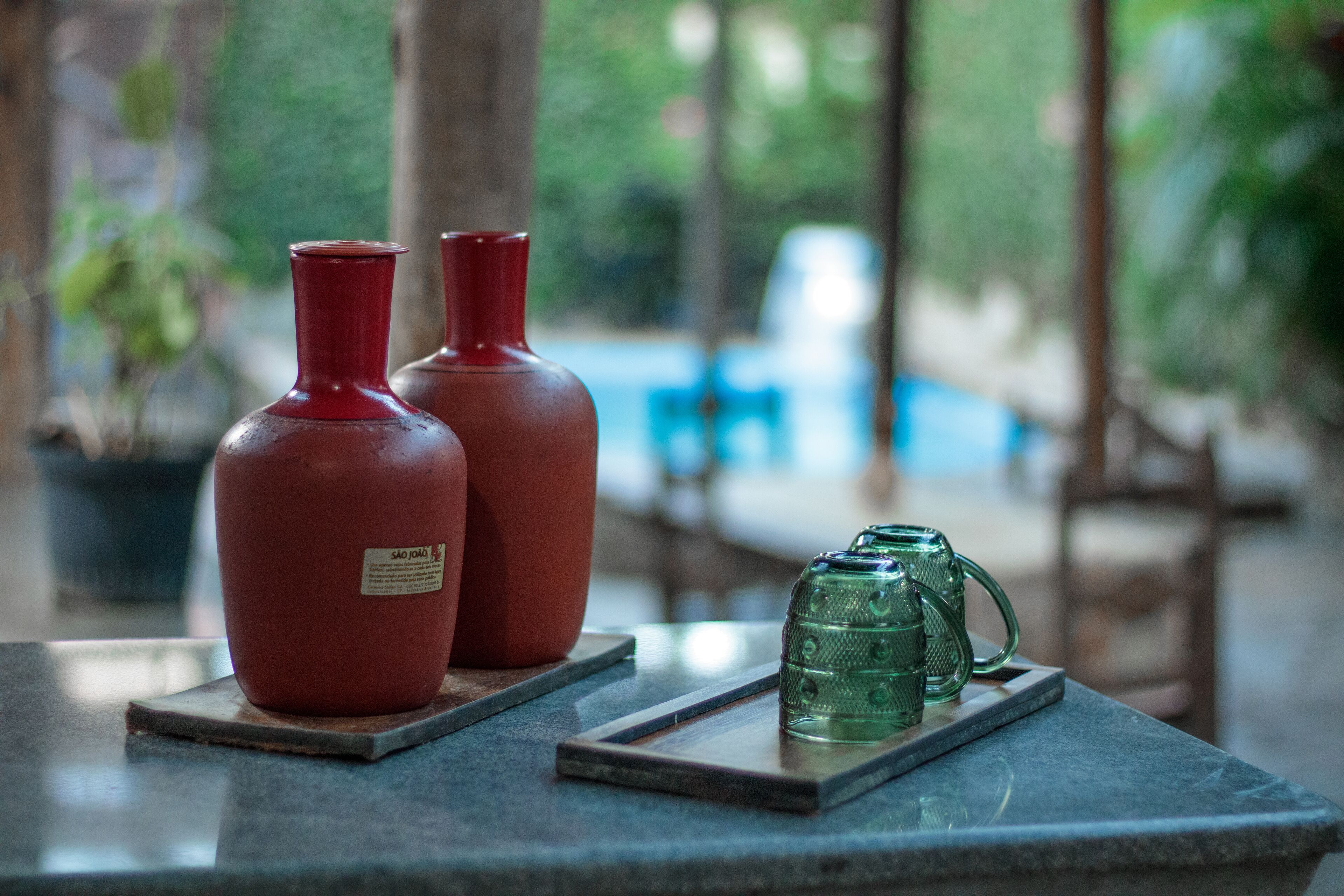 Two red bottles and a green glass mug on a table with a view of a pool and greenery in the background.
