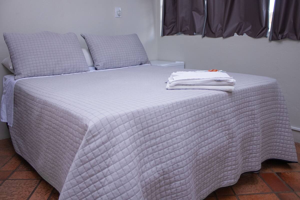 A neatly made bed with a quilted bedspread and pillows, a folded white towel on the bed, and a window with curtains.