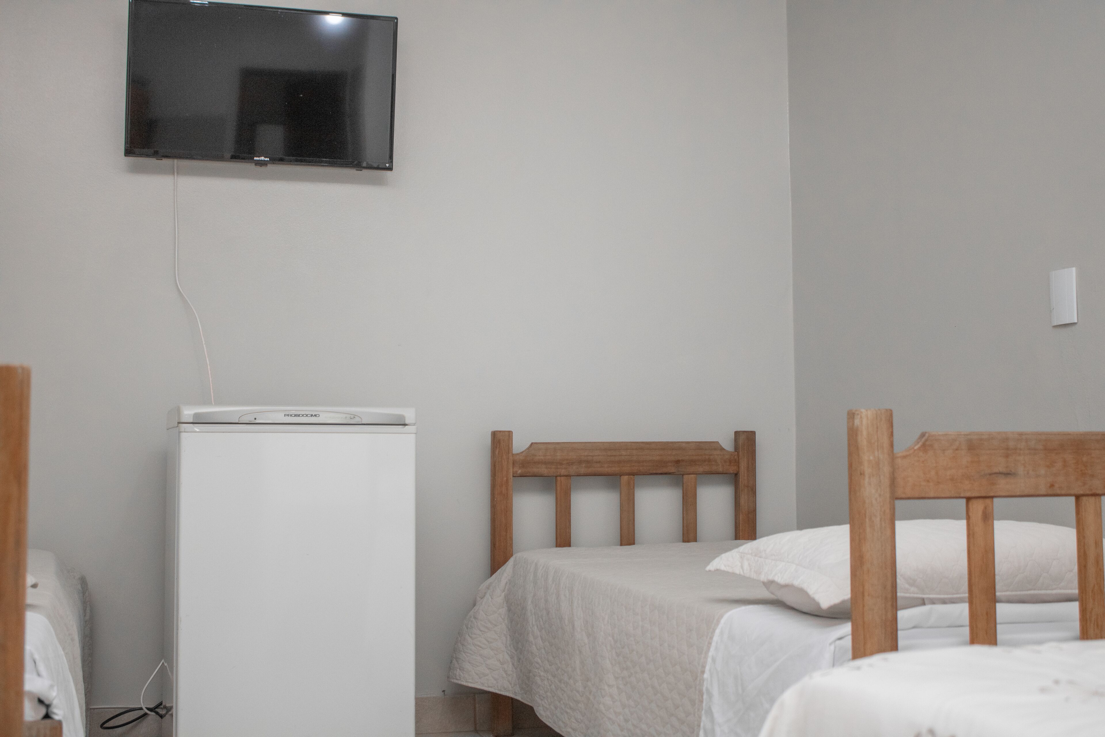 Un dormitorio con cama, cabecero de madera, mesita de noche blanca y televisor instalado en la pared.