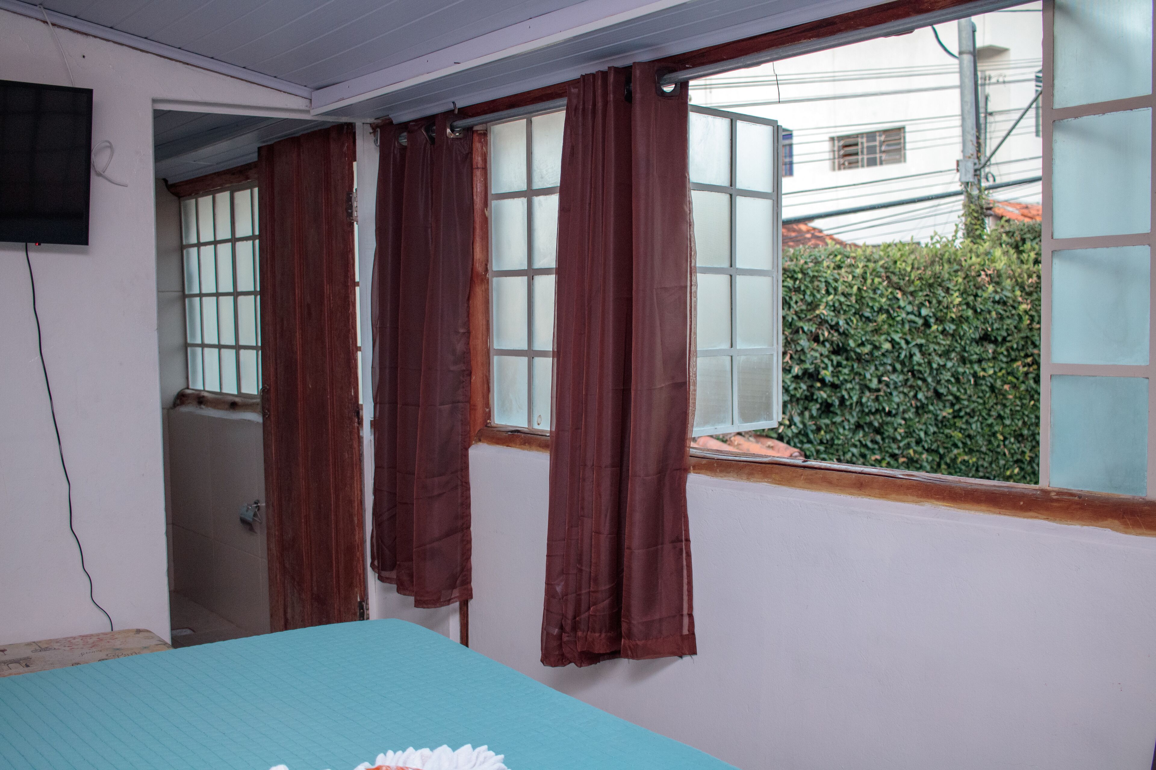 Una habitación con cama, ventana con cortinas, televisor en la pared y vistas a un edificio y zonas verdes.