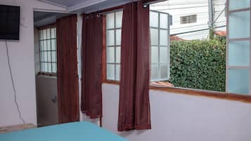 Una habitación con cama, ventana con cortinas, televisor en la pared y vistas a un edificio y zonas verdes.