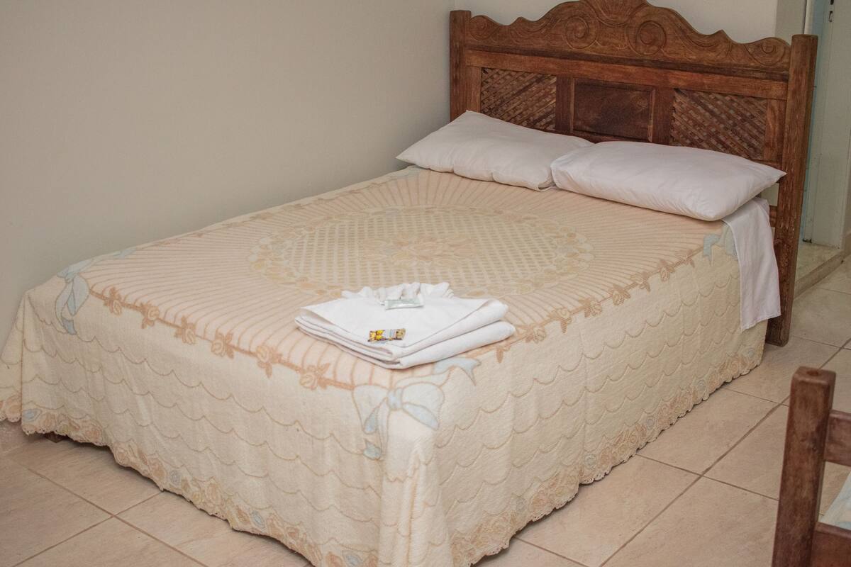A neatly made bed with a patterned bedspread and pillows, a wooden headboard, and a small bedside table with a folded towel and a bottle.