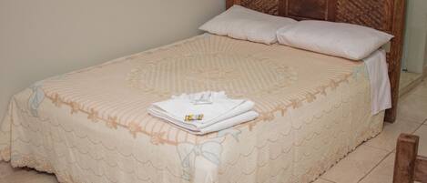 A neatly made bed with a patterned bedspread and pillows, a wooden headboard, and a small bedside table with a folded towel and a bottle.