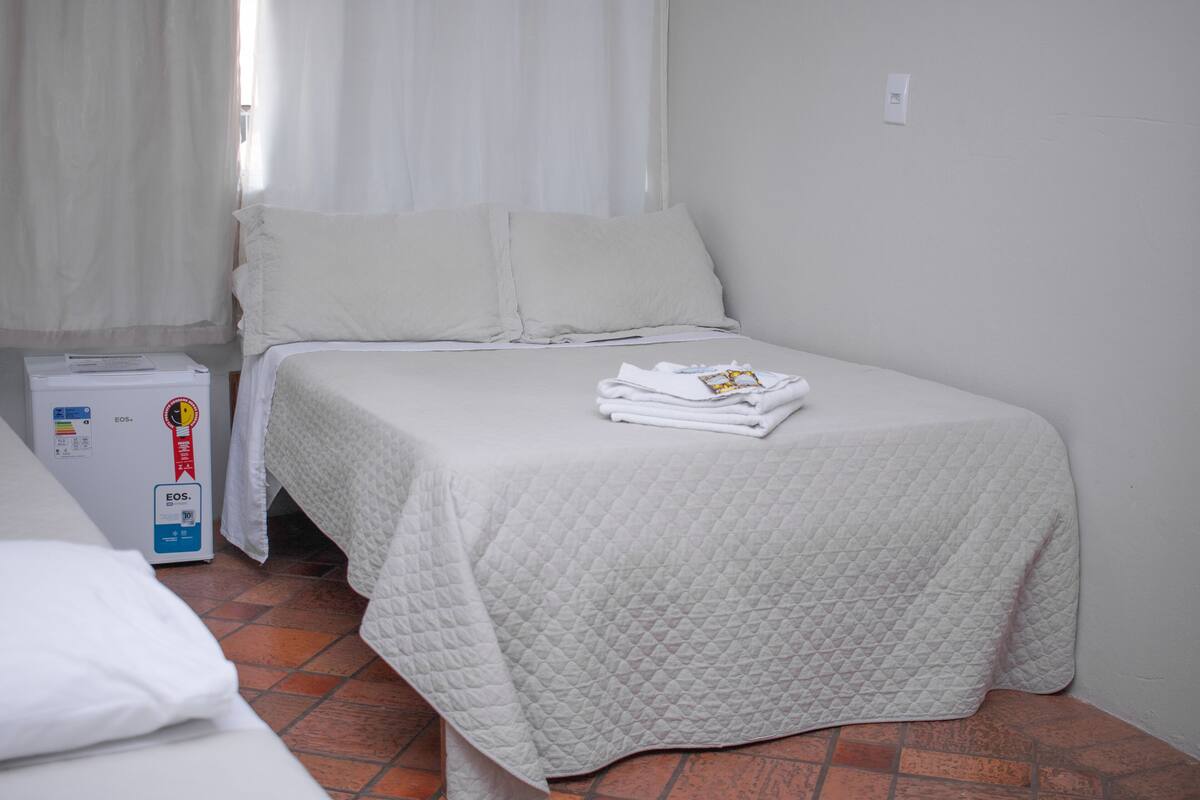 A single bed with a quilted white bedspread, a white pillow, and a folded towel on the bed. There is a white heater unit beside the bed and a white wall with a light switch.
