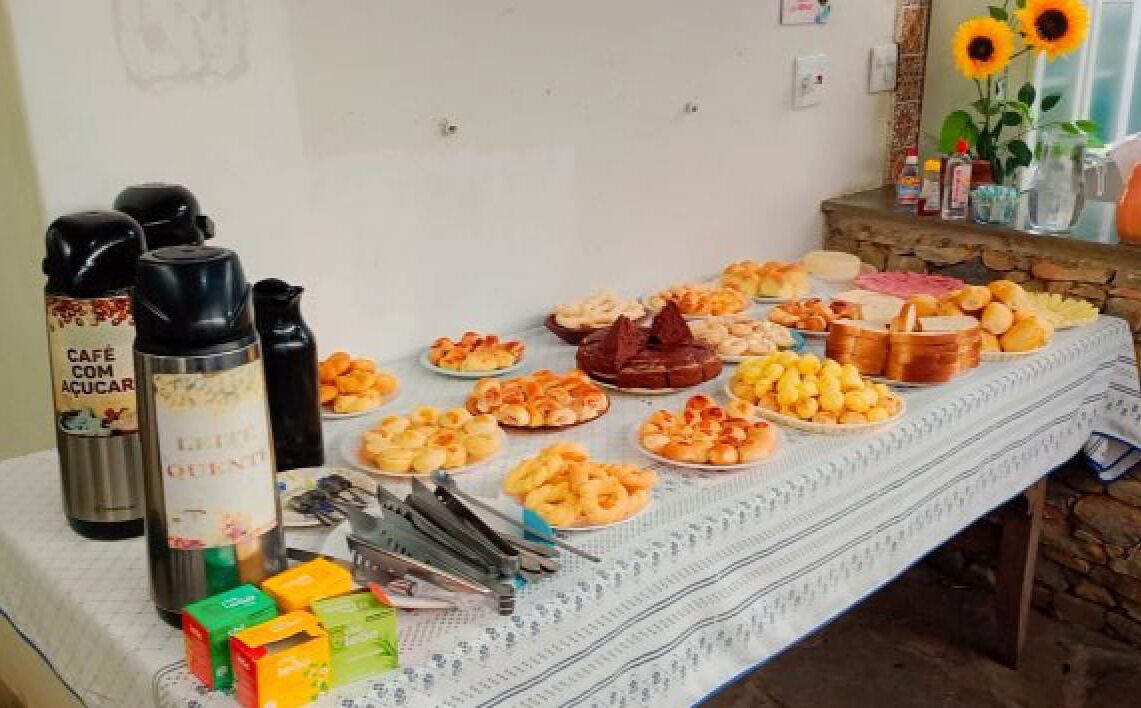 Uma mesa de buffet com diversos alimentos, acompanhamentos, uma cafeteira e um jarro de água.