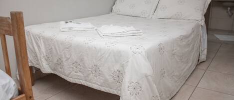 Un lit bien fait, recouvert d’une couvre-lit à motifs floraux et agrémenté d’oreillers, une chaise en bois et un sol carrelé.