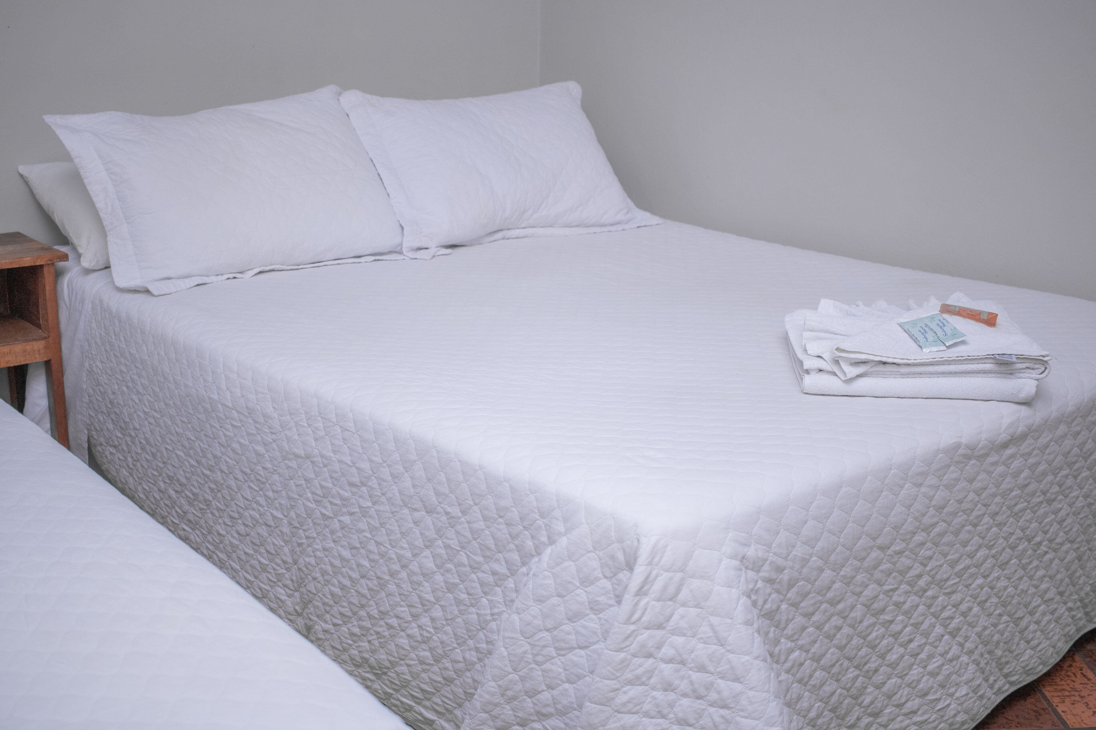 A neatly made bed with white bedding and pillows, a wooden nightstand, and a folded towel on the bed.