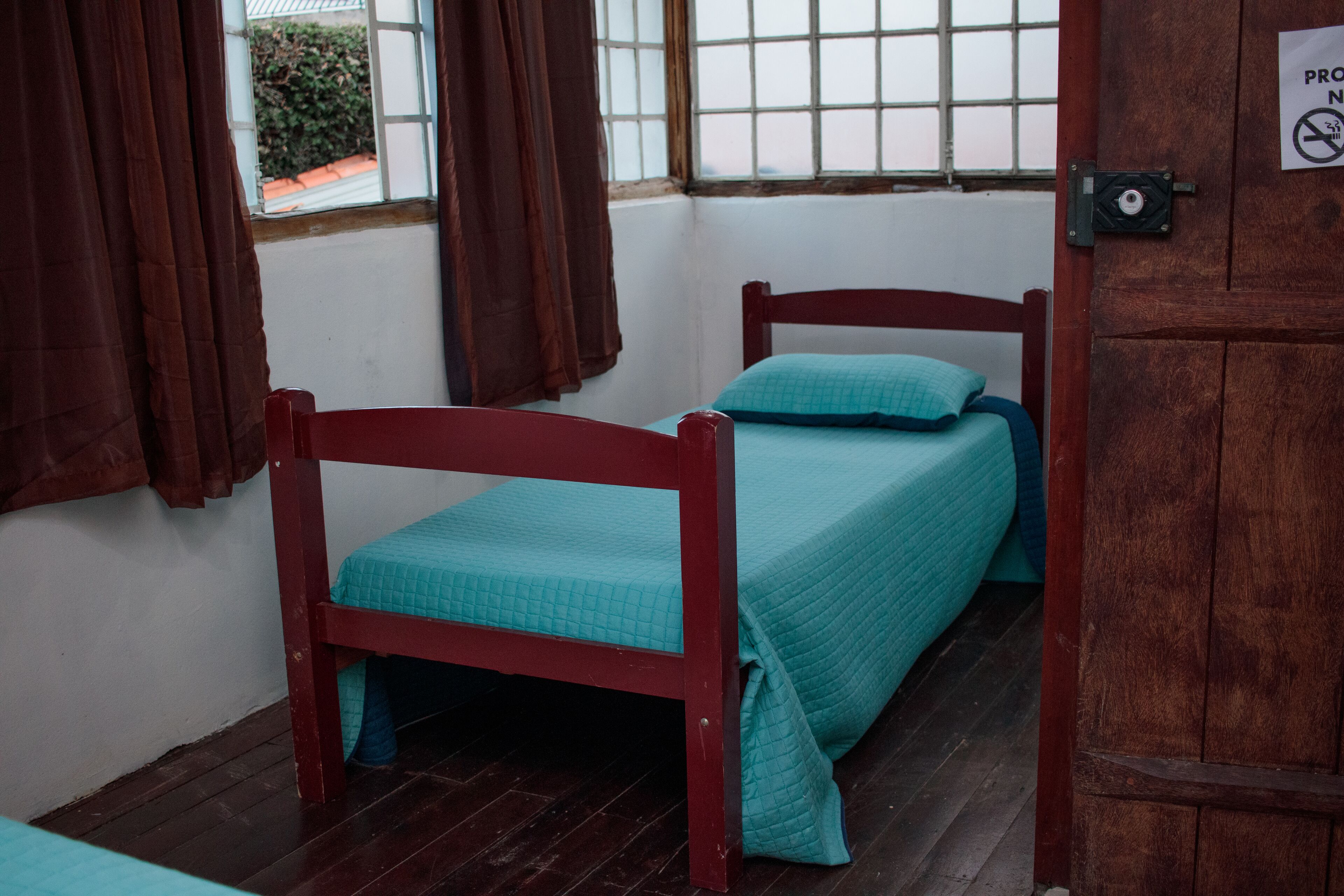 A single bed with a wooden frame and teal bedding in a room with a wooden floor and a window with brown curtains.