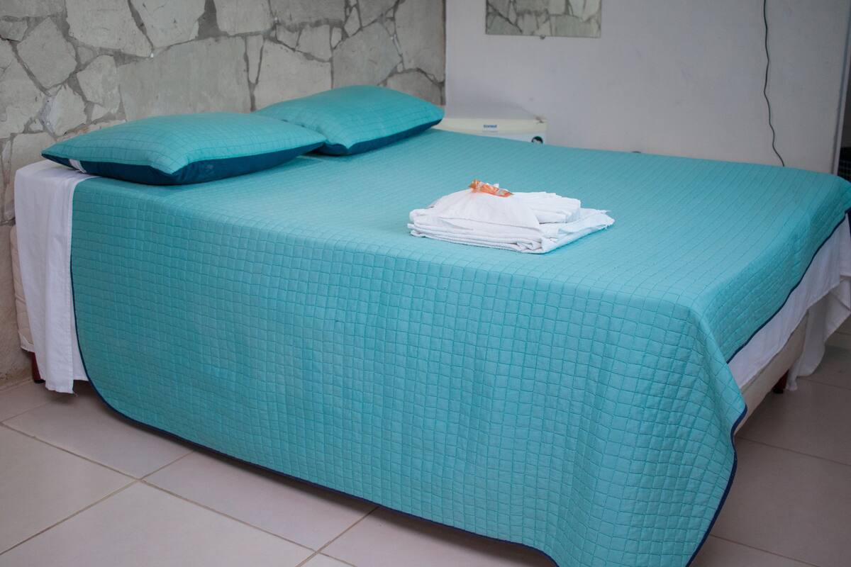 A bed with a teal quilted bedspread, a white pillow, and a neatly folded white towel on the bed.