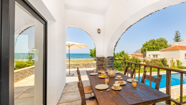 A wooden dining table set with plates, cups, and a glass of orange juice, overlooking a pool and the sea. - Platanias Beachfront Villa Elsa-90m to main Square (GW95+FMM Platanias, Greece)