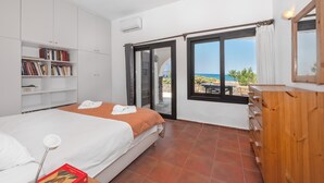 A bedroom with a bed, a wooden dresser, a bookshelf, and a view of the sea through a large window. - Platanias Beachfront Villa Elsa-90m to main Square (GW95+FMM Platanias, Greece)