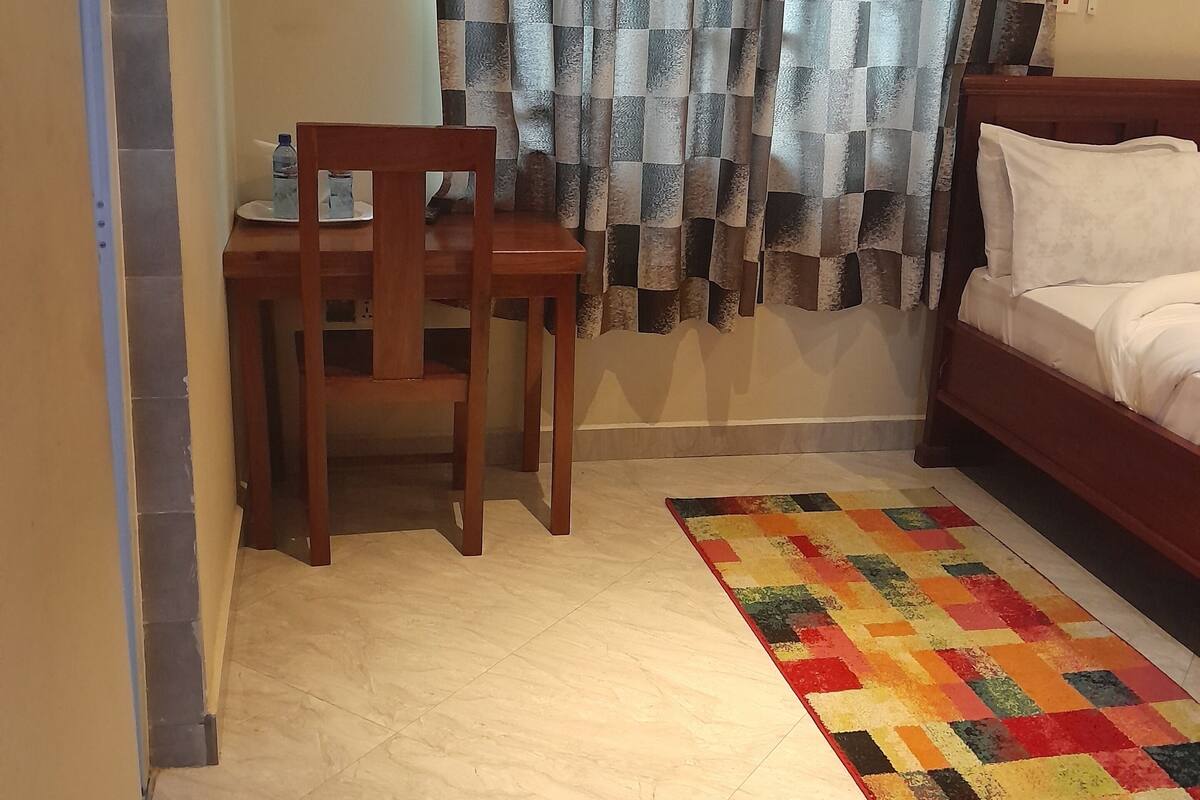 Ein Schlafzimmer mit einem Bett, einem Holzstuhl, einem kleinen Tisch mit einer Wasserflasche, einem farbenfrohen Teppich und einem karierten Vorhang.
