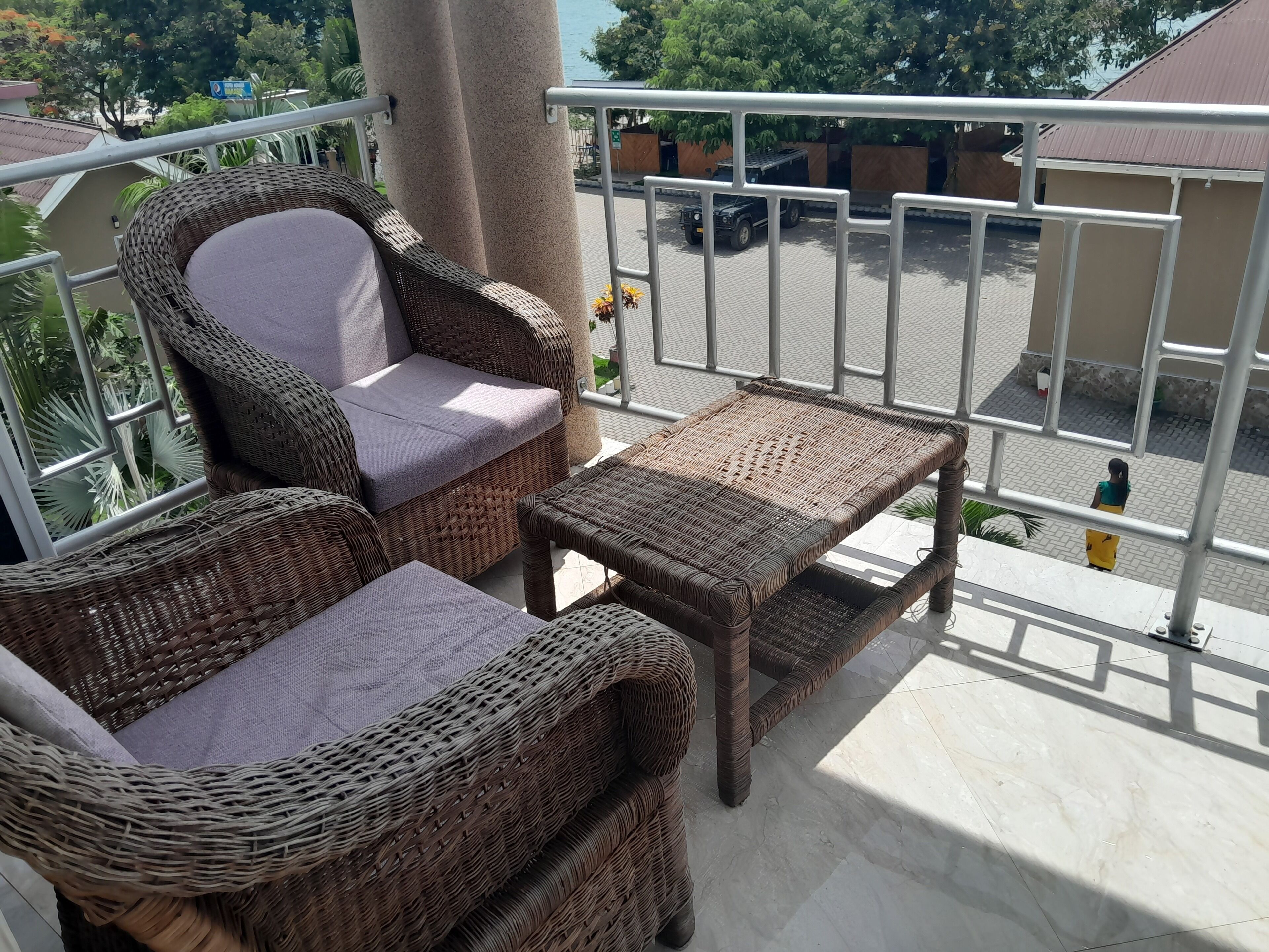 Un balcon doté de meubles en osier, d’une rambarde en métal et offrant une vue sur une rue où est garé un véhicule et où une personne se promène.
