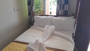 Ein ordentlich bezogenes Bett mit weißen Leinen und einem braunen Kopfteil, ein Fenster mit Vorhängen und ein an der Wand befestigter Lichtschalter.