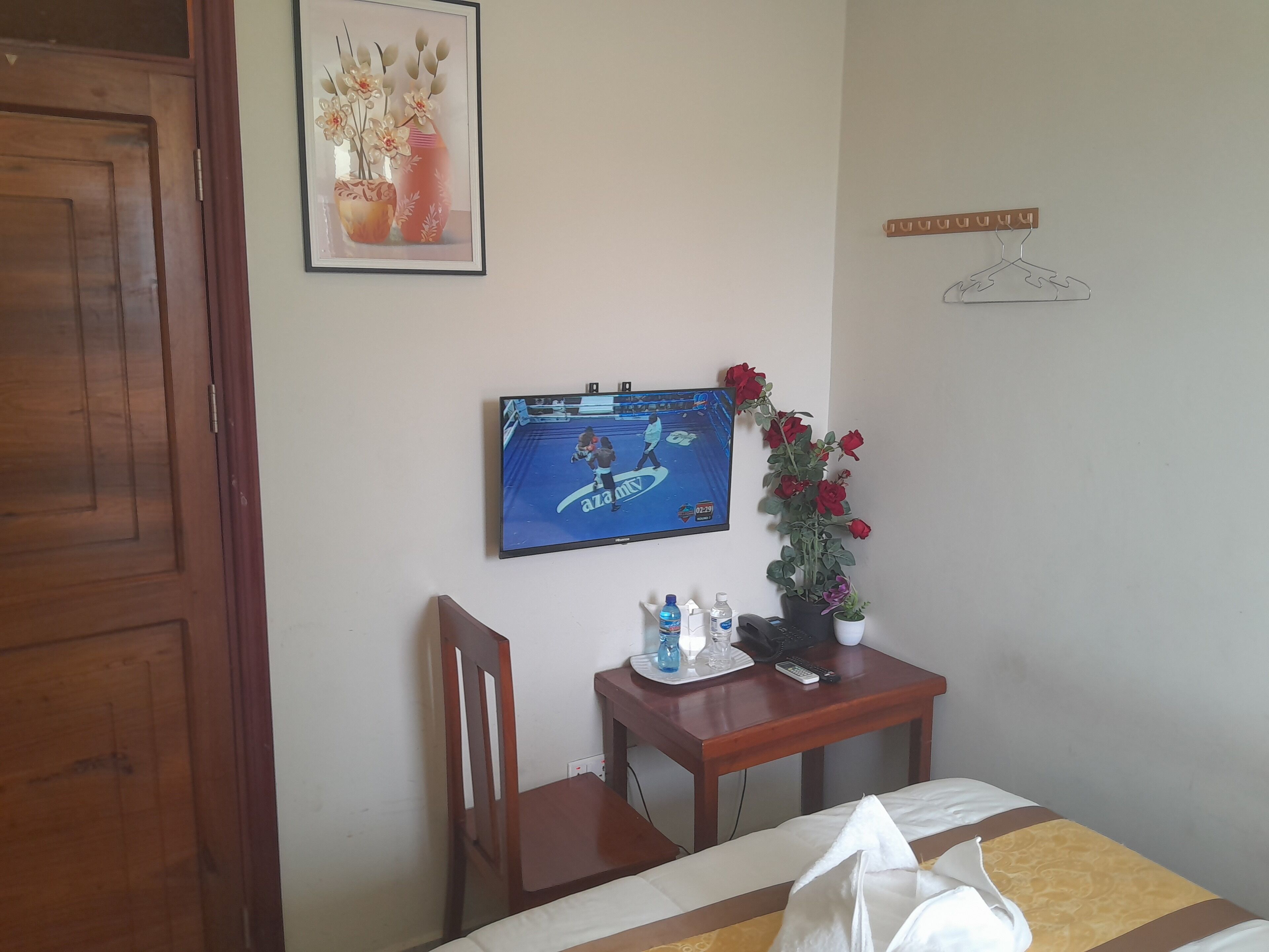 A hotel room with a bed, a desk with a TV, a chair, a small table with a vase of flowers, and a wall-mounted hanger.