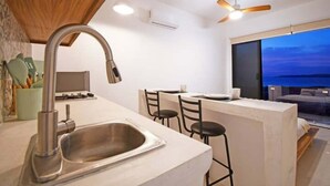 Una cocina moderna con un lavamanos de acero inoxidable, una zona de comedor con taburetes de bar y un amplio ventanal con vistas al océano al atardecer.