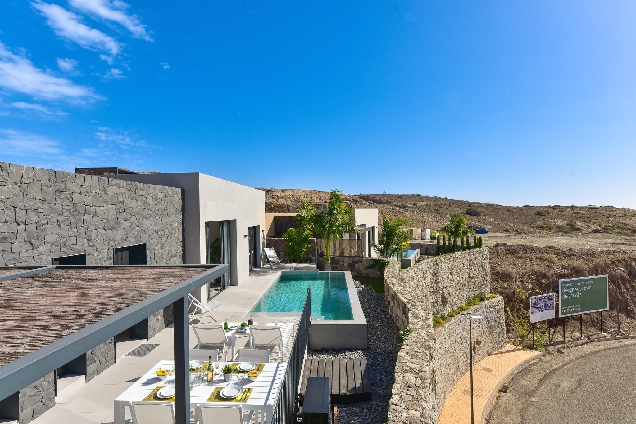 A modern outdoor pool area with a stone wall, a dining setup, and a view of the surrounding landscape.