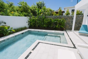 A swimming pool with a stone wall and tiled deck, surrounded by lush greenery and a white building. - Pearl Villa (Grand-Baie)