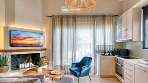 A modern living room with a fireplace, a flat-screen TV, a blue armchair, and a kitchen area with cabinets and a microwave. - Deluxe Seaside Condo for 6 (Kantia)