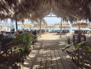 En restaurang vid stranden med uteservering, parasoller med halmtak och en träbrygga som leder ner till stranden.