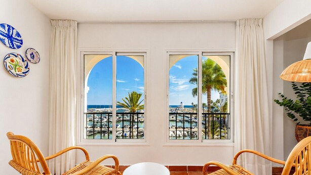 Una habitación con dos sillones de mimbre, una mesita blanca y vistas a un puerto deportivo a través de amplias ventanas.